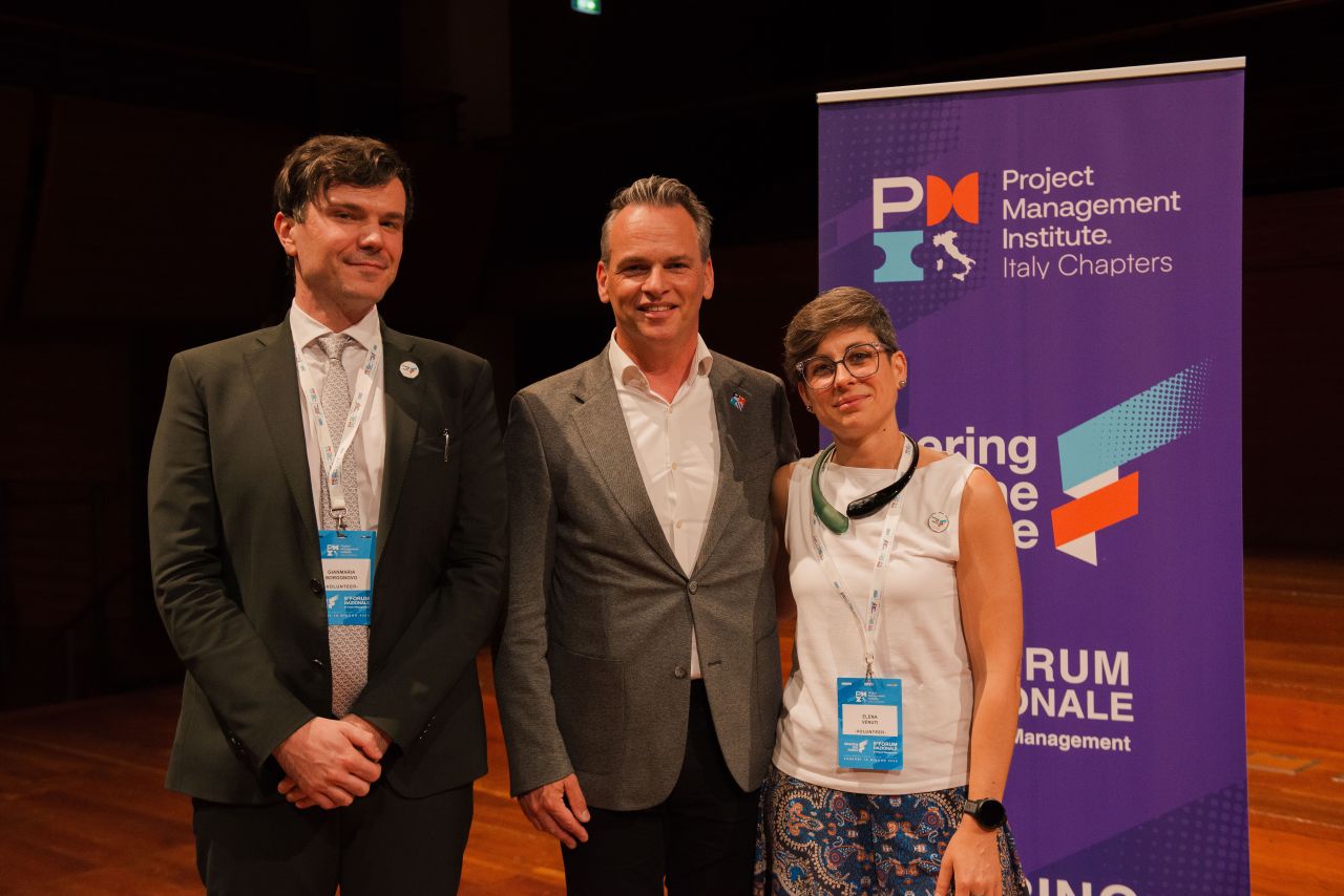 1781944717773 Three professionals pose on a stage beside a purple Project Management Institute banner showing Italy Chapters signage, smiling at the camera as a formal event backdrop.