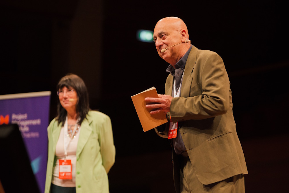 DSC_0897 Bald man in a tan suit on stage with a notebook and headset mic, speaking at a conference; woman in a mint blazer stands behind him.